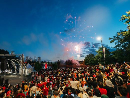 A large crowd gathers at an outdoor event during twilight to watch fireworks, with city buildings in